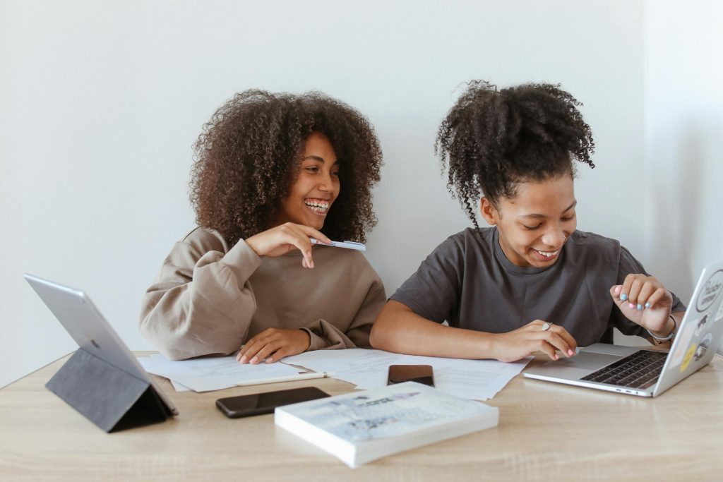 Two teenage girls studying together at a desk with laptops, smiling and focused.