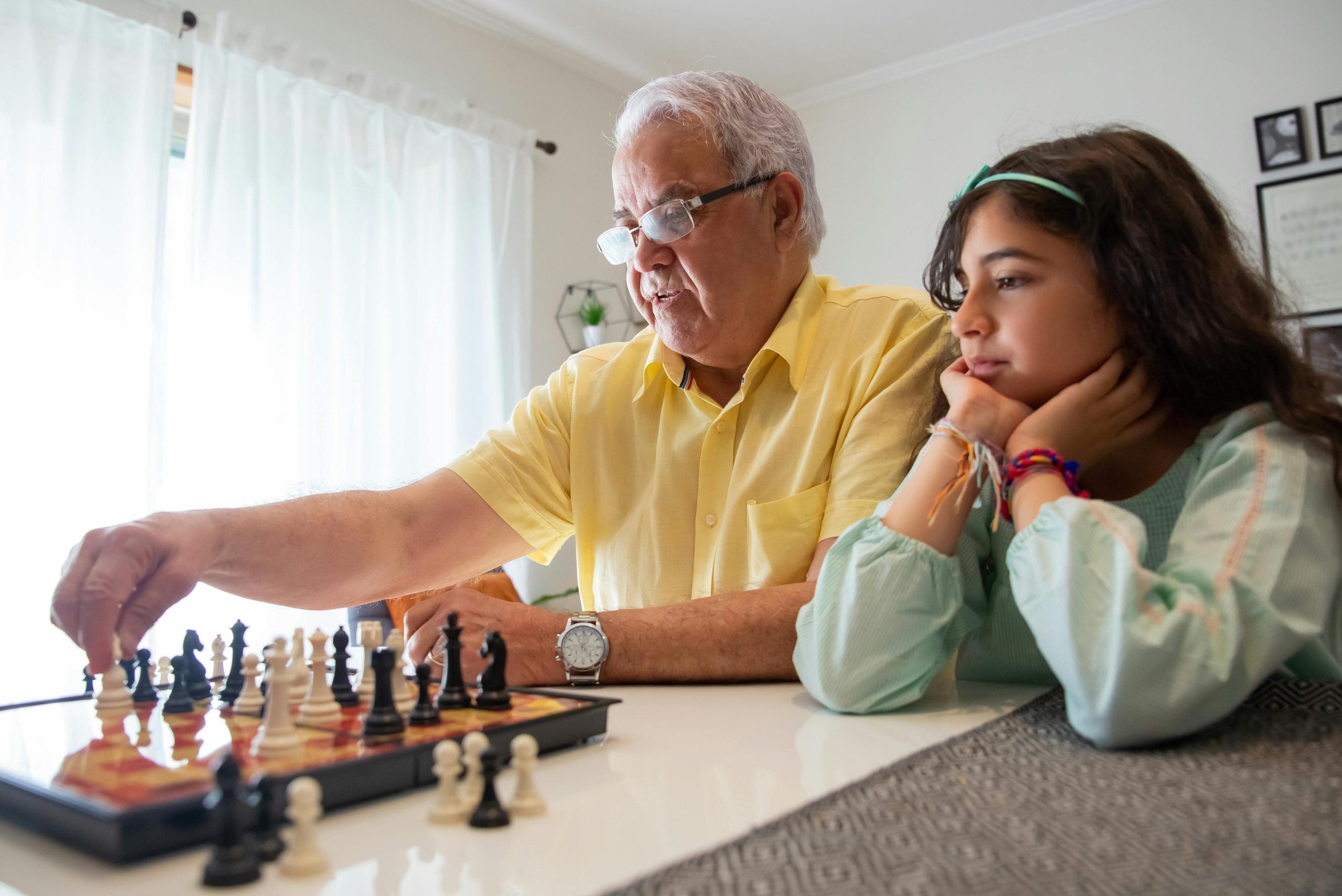 Elderly man and young girl bond over a chess game indoors, capturing a moment of family connection.
